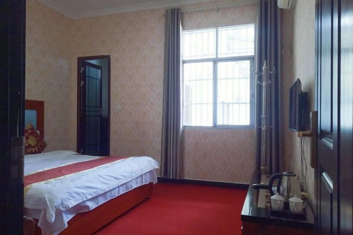 A hotel room with a bed, a window with curtains, a bedside table with cups, and a television on the wall.