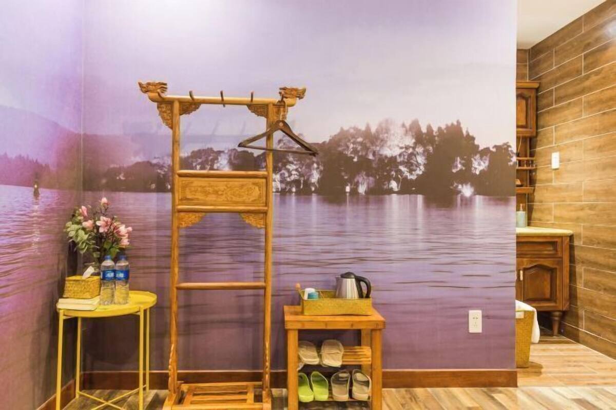 Een kamer met houten vloer, een houten tafel met een vaas en flessen, een houten stoel, een houten plank met schoenen, en een muurschildering van een meer.
