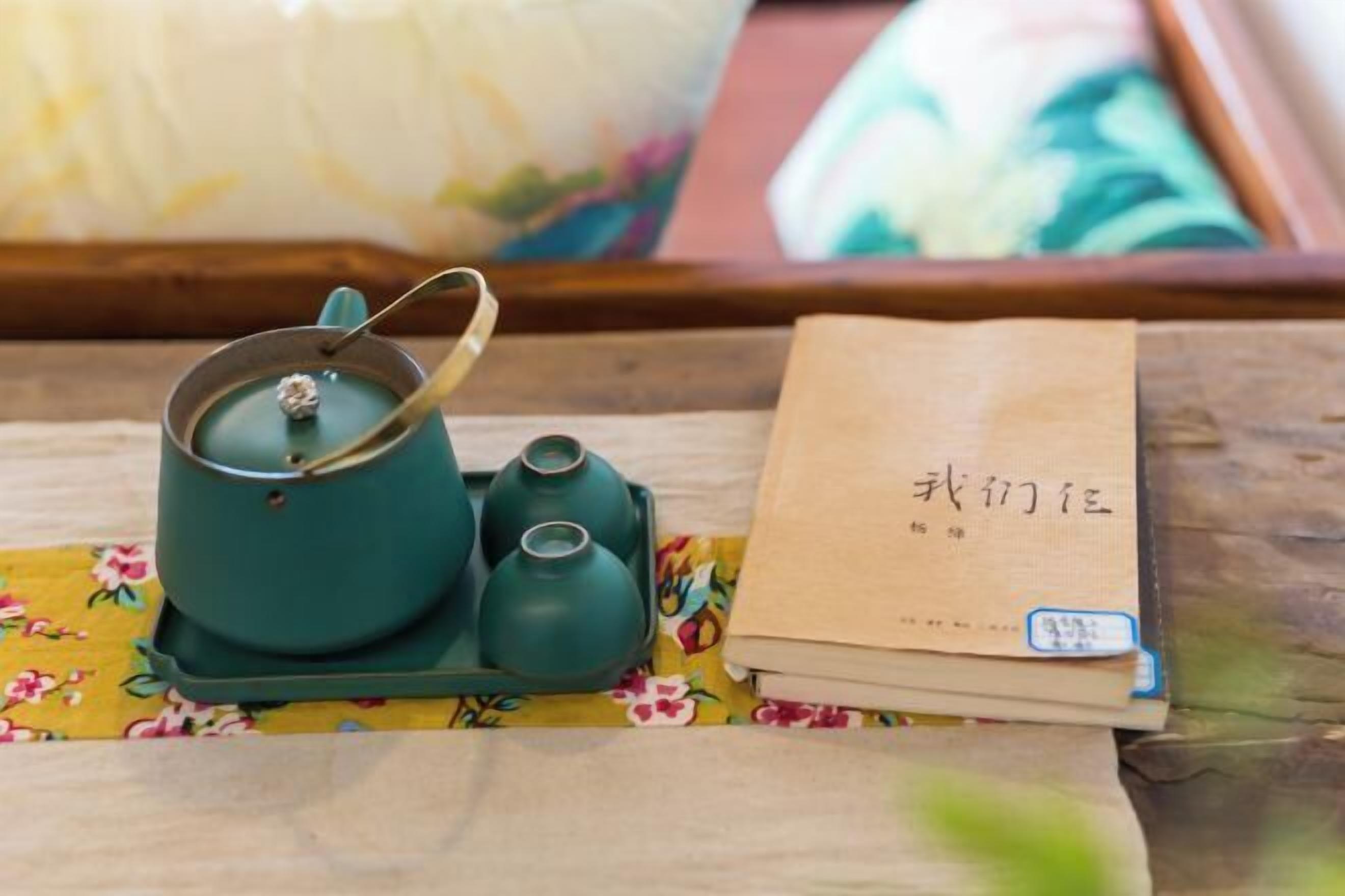 Una bandeja de madera con una tetera verde azulada, dos tazas de té pequeñas y un libro marrón con caracteres chinos.