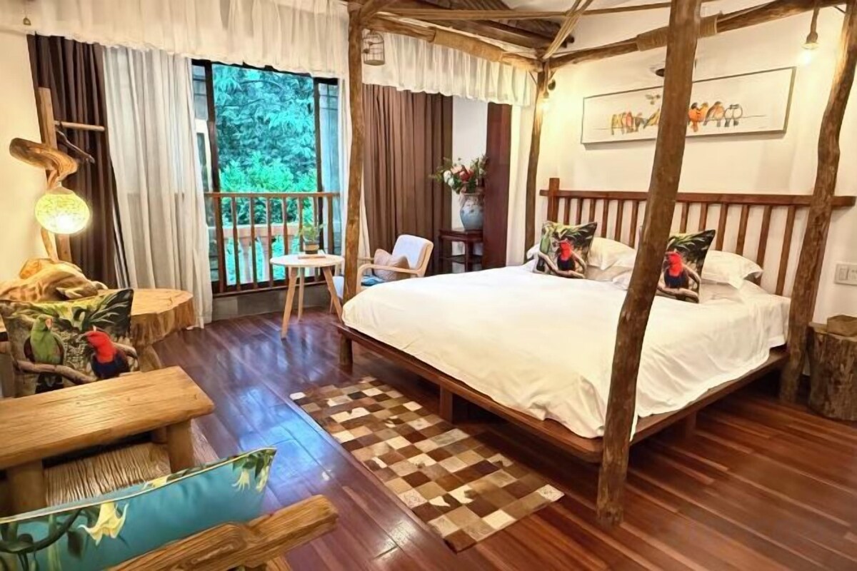 A bedroom with a large bed, wooden flooring, a small table, and a view of the outdoors.