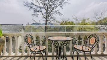 A balcony with two metal chairs and a table, a glass railing, and a view of trees and buildings.