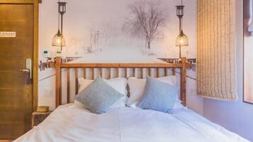 Una cama bien hecha con cabecera de madera, dos lámparas de noche y un cuadro en la pared que representa un paisaje nevado.