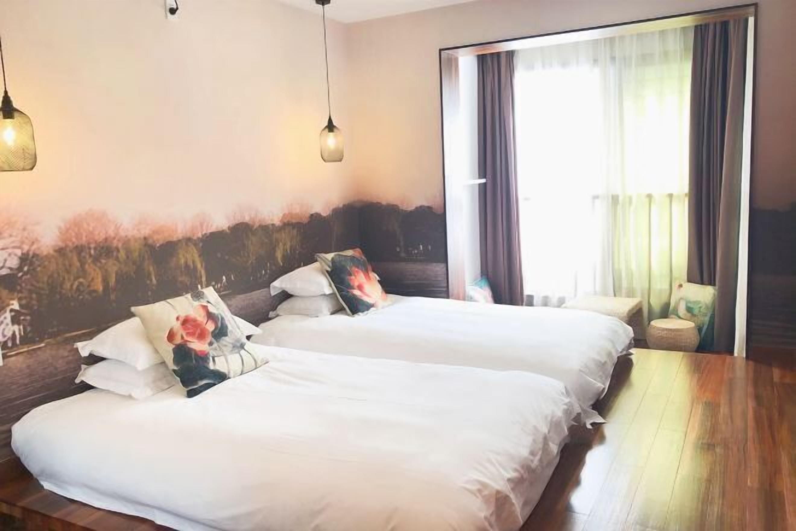 A hotel room with two beds, a wooden floor, a large window with curtains, and wall-mounted lights.