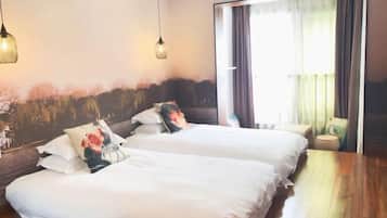 A hotel room with two beds, a wooden floor, a large window with curtains, and wall-mounted lights.