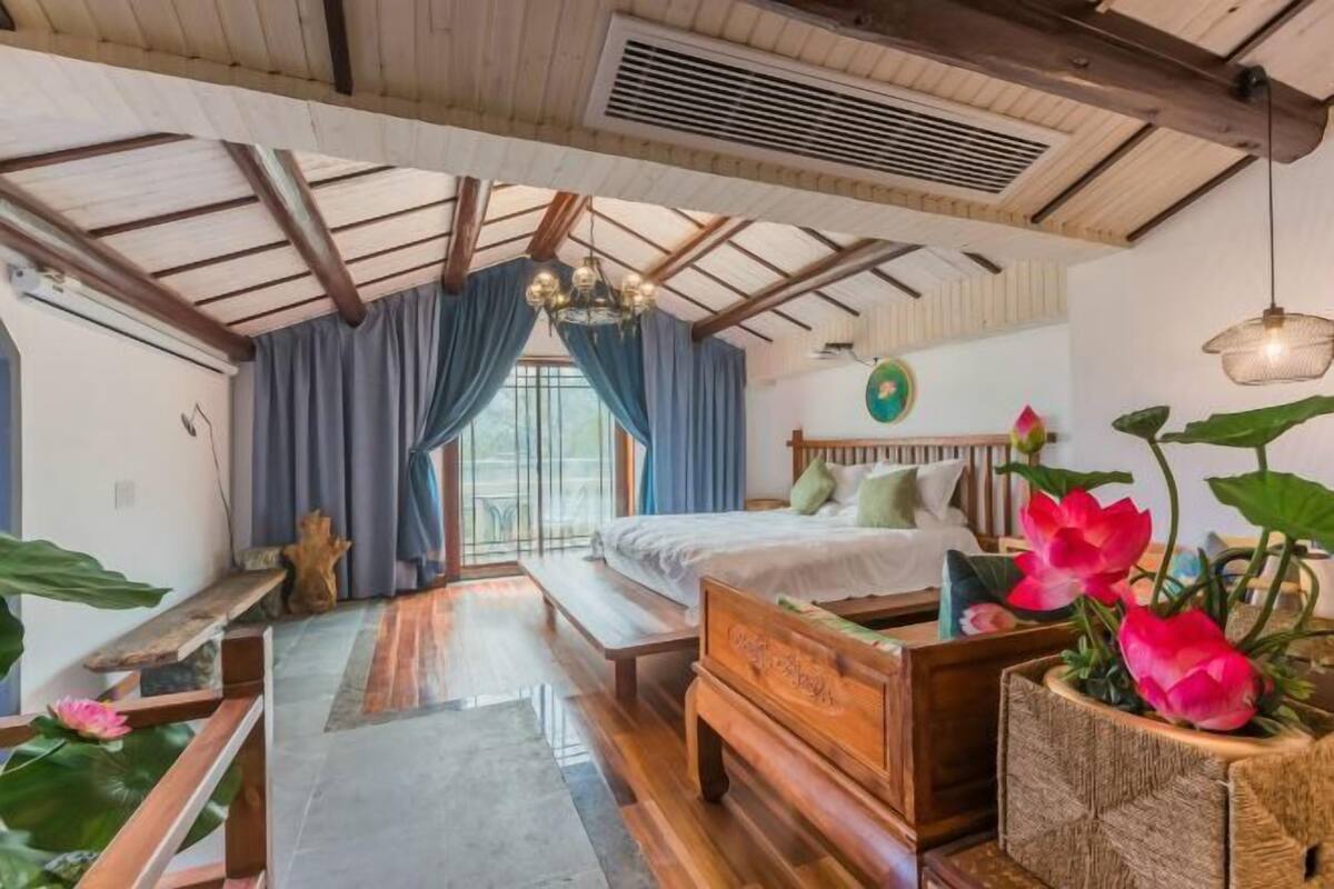 Een slaapkamer met houten plafond, een bed met kussens, een bankje, een raam met gordijnen en een hanglamp.
