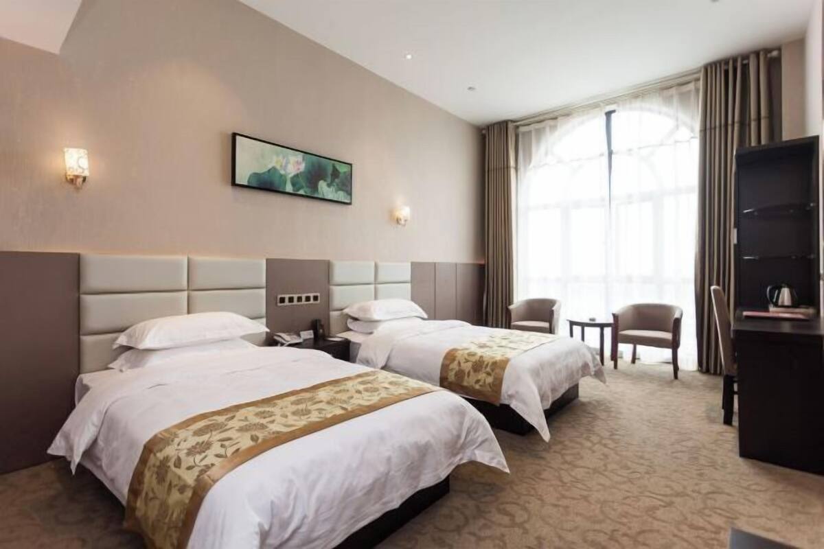 A hotel room with two beds, a desk, a chair, a TV, and a large window with curtains.