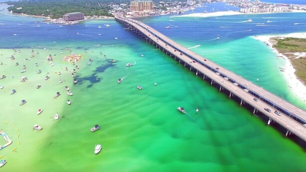 A coastal bridge with boats in the water, a beach, and a cityscape in the background.