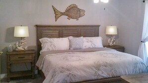 A bedroom with a bed, two bedside tables, a nightstand, a fish wall decoration, and a window with curtains.