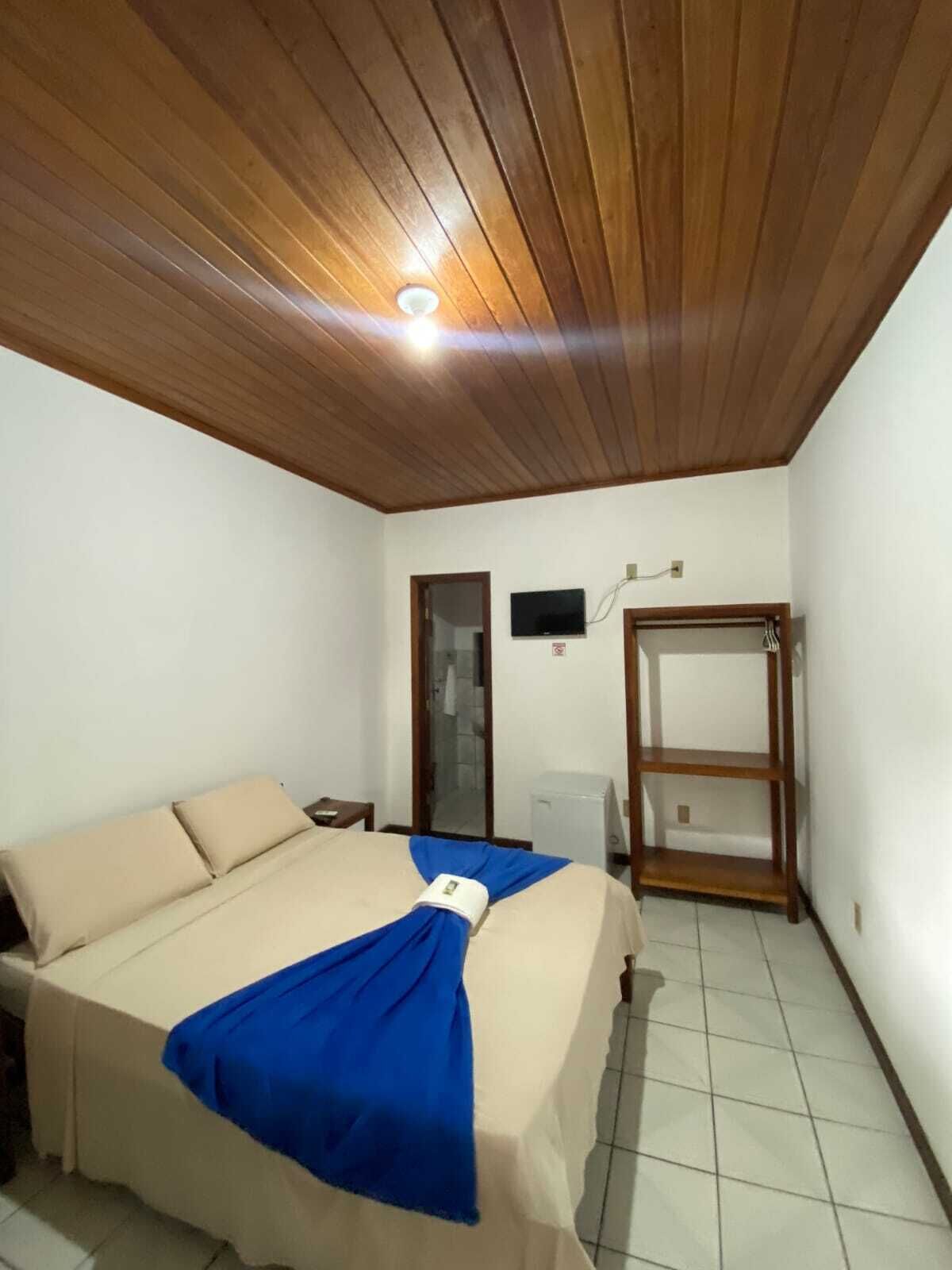 A bedroom with a wooden ceiling, a bed with a blue blanket, a TV on the wall, and a wooden wardrobe.