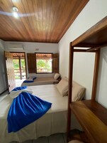 Una habitación con techo de madera, cama con manta azul, ventana con vista a la vegetación y un banco de madera.