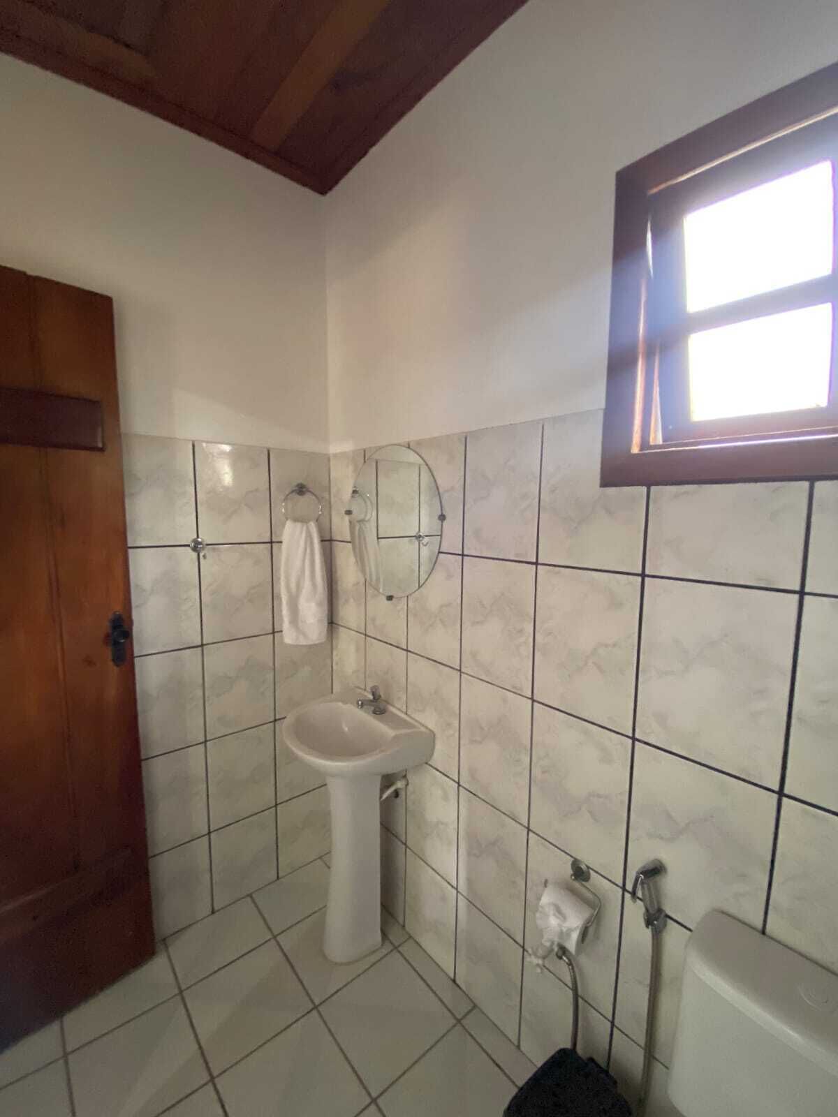 Casa de banho com lavatório branco, espelho e sanita. Porta de madeira e janela com vista para o exterior.