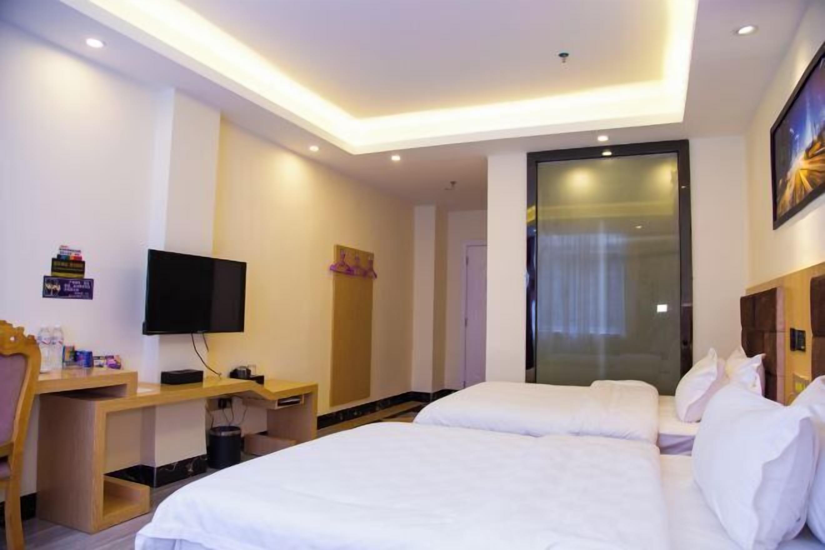 A hotel room with two beds, a desk with a flat-screen TV, a wooden nightstand, and a glass door leading to a bathroom.