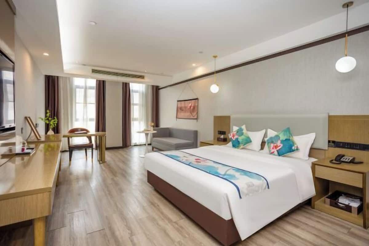 A hotel room with a large bed, a desk, a chair, a sofa, and a bedside table with a lamp.