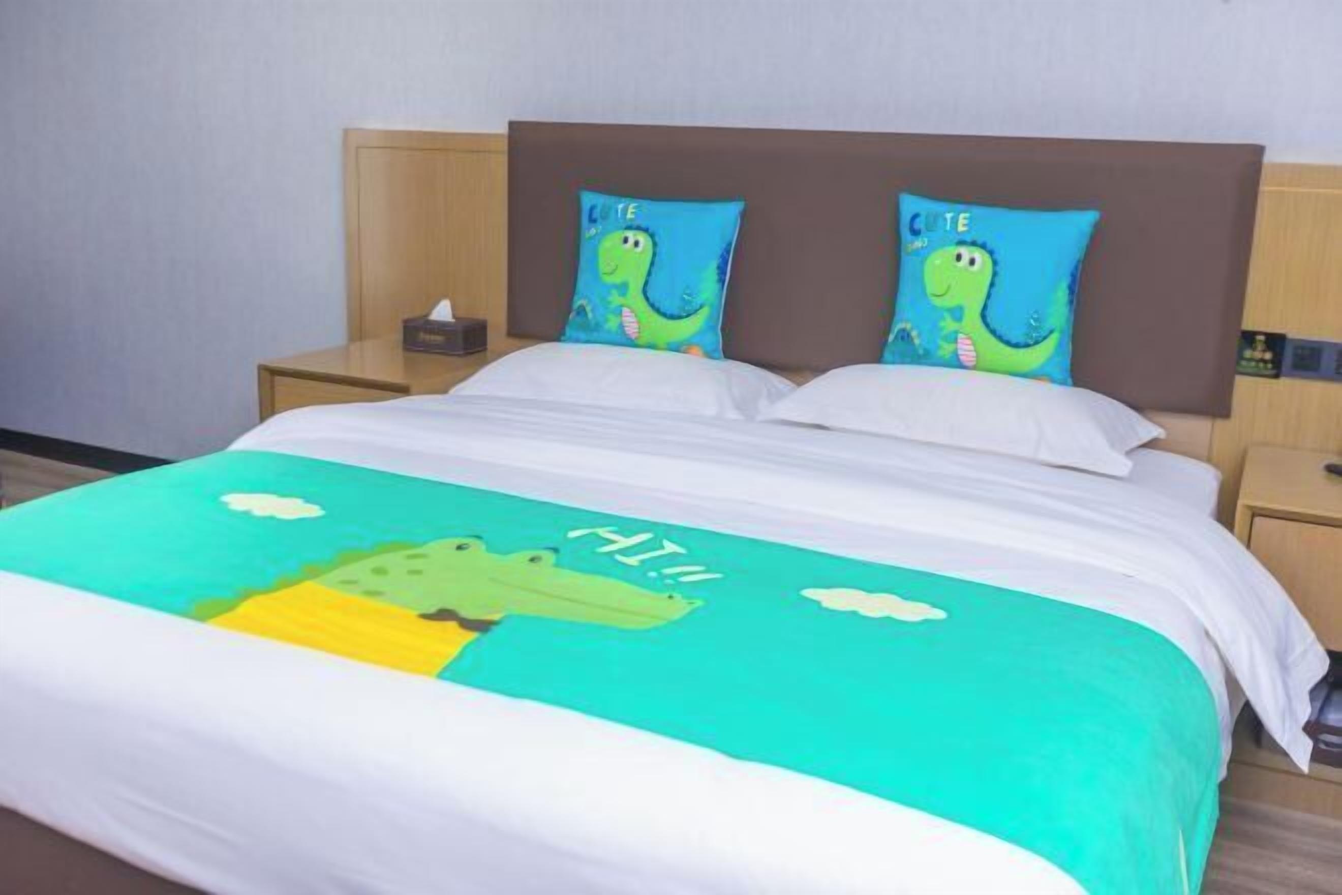 Un lit avec une couvre-lit verte et blanche ornée d’un crocodile et de nuages, deux oreillers à motifs de dinosaures et une tête de lit en bois.