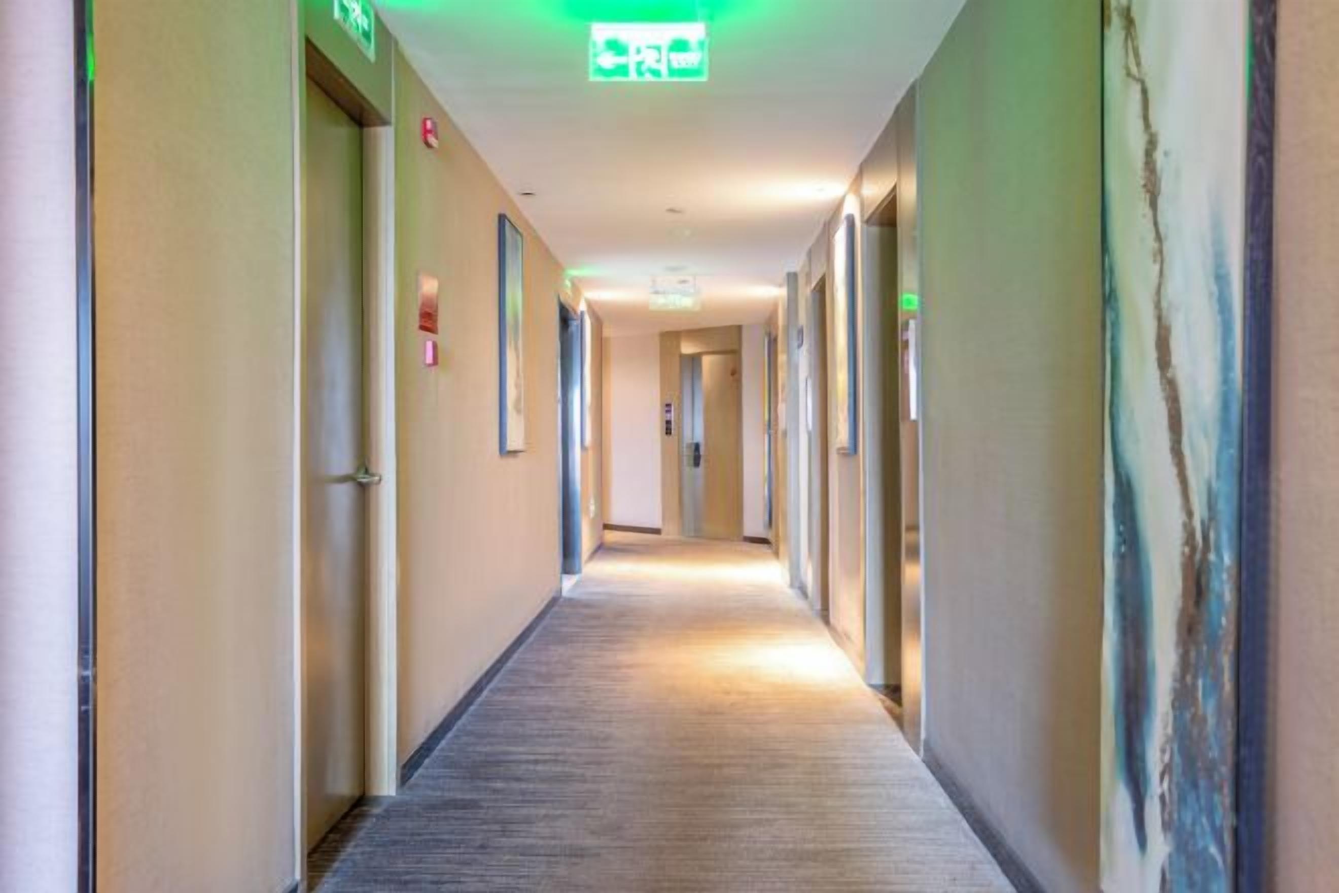 Un pasillo de hotel con puertas a ambos lados, suelo cubierto de alfombra y un letrero de salida verde en el techo.