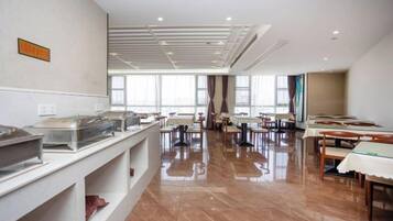 Un comedor amplio con mesas y sillas, una barra de buffet con recipientes de comida, y ventanas grandes que dejan entrar luz natural.