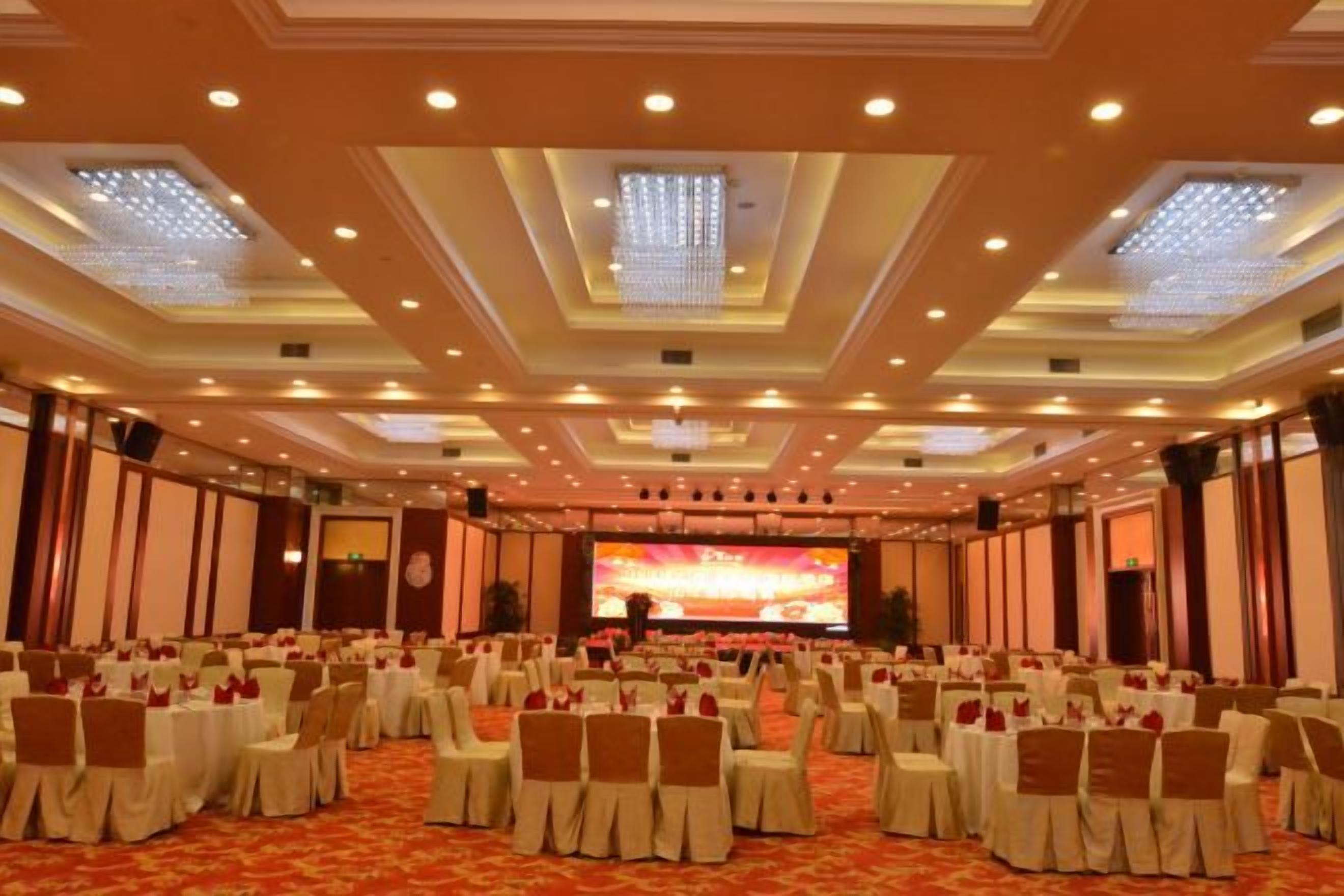 Un salón de conferencias amplio con filas de sillas y mesas, un escenario con una pantalla grande e iluminación empotrada en el techo.