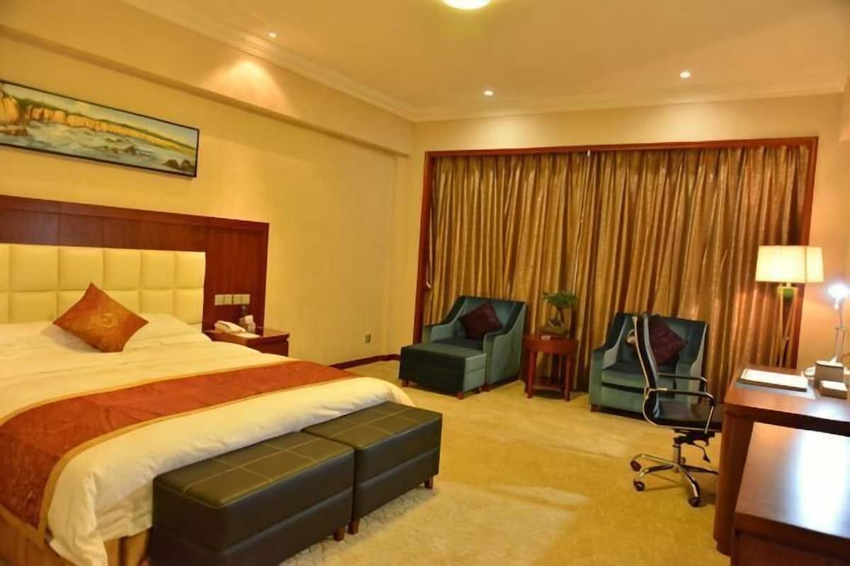 Une chambre d’hôtel avec un grand lit, deux fauteuils, un bureau et une grande fenêtre avec des rideaux.