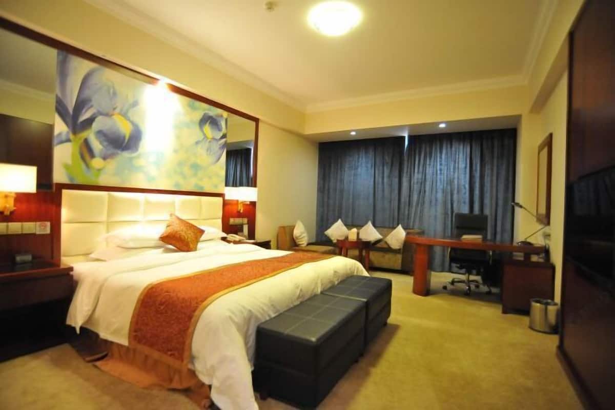 Une chambre d’hôtel avec un grand lit, un bureau, une chaise, un banc, une télévision et un tableau au mur.
