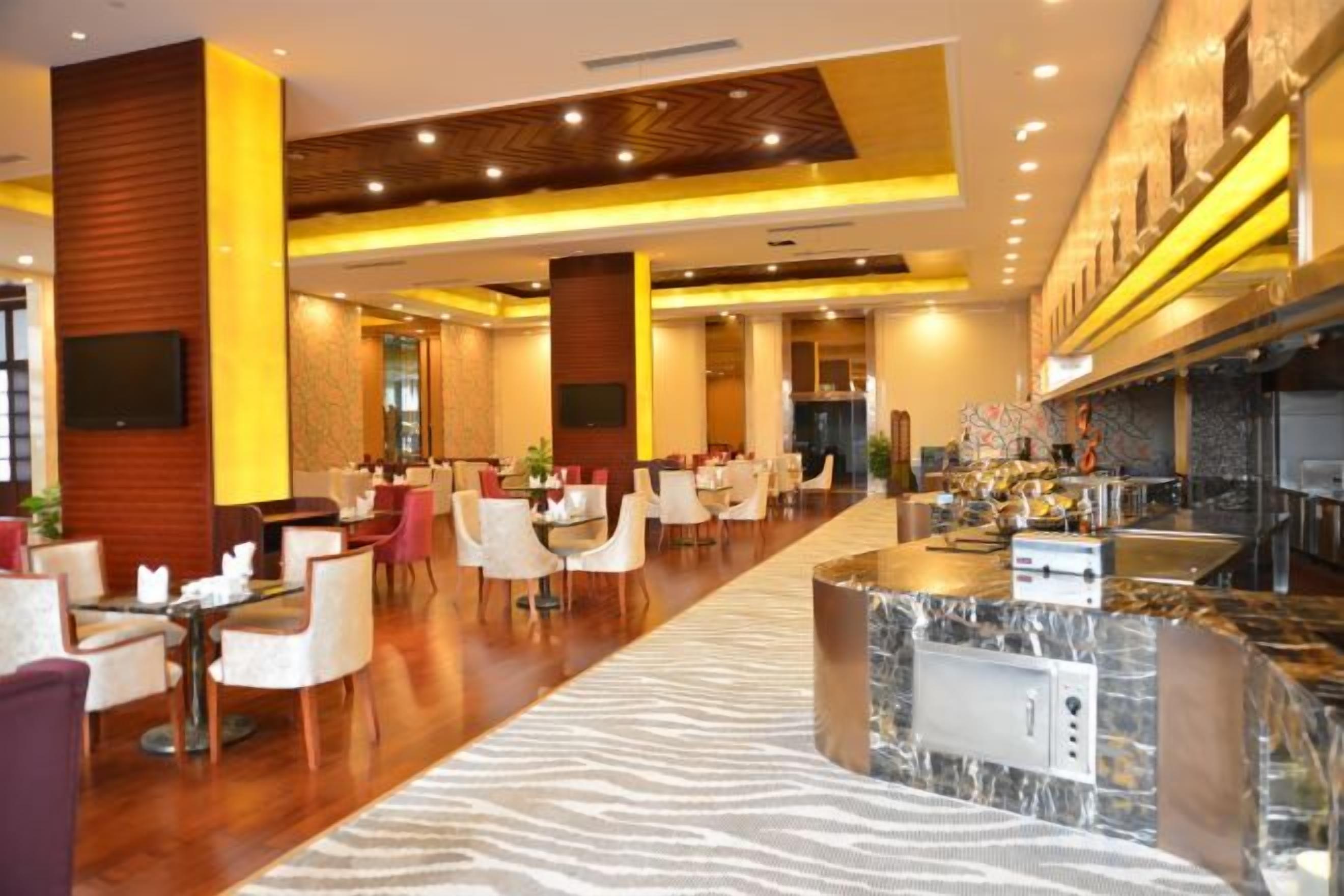 A hotel lobby with a long reception desk, multiple seating areas, and a modern kitchen visible in the background.