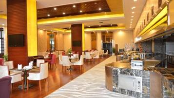 A hotel lobby with a long reception desk, multiple seating areas, and a modern kitchen visible in the background.