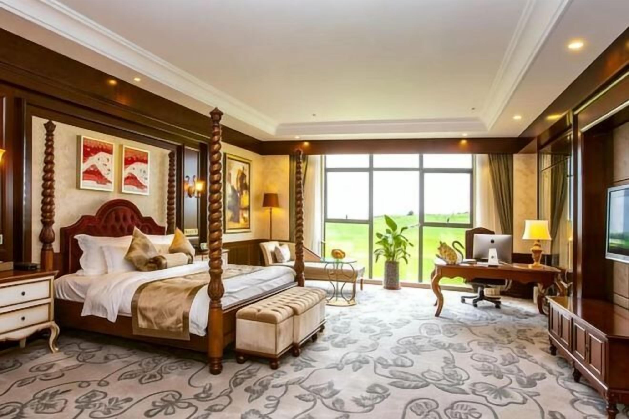 A four-poster bed with a tufted headboard, a desk with a computer, and a large window with a view of greenery.