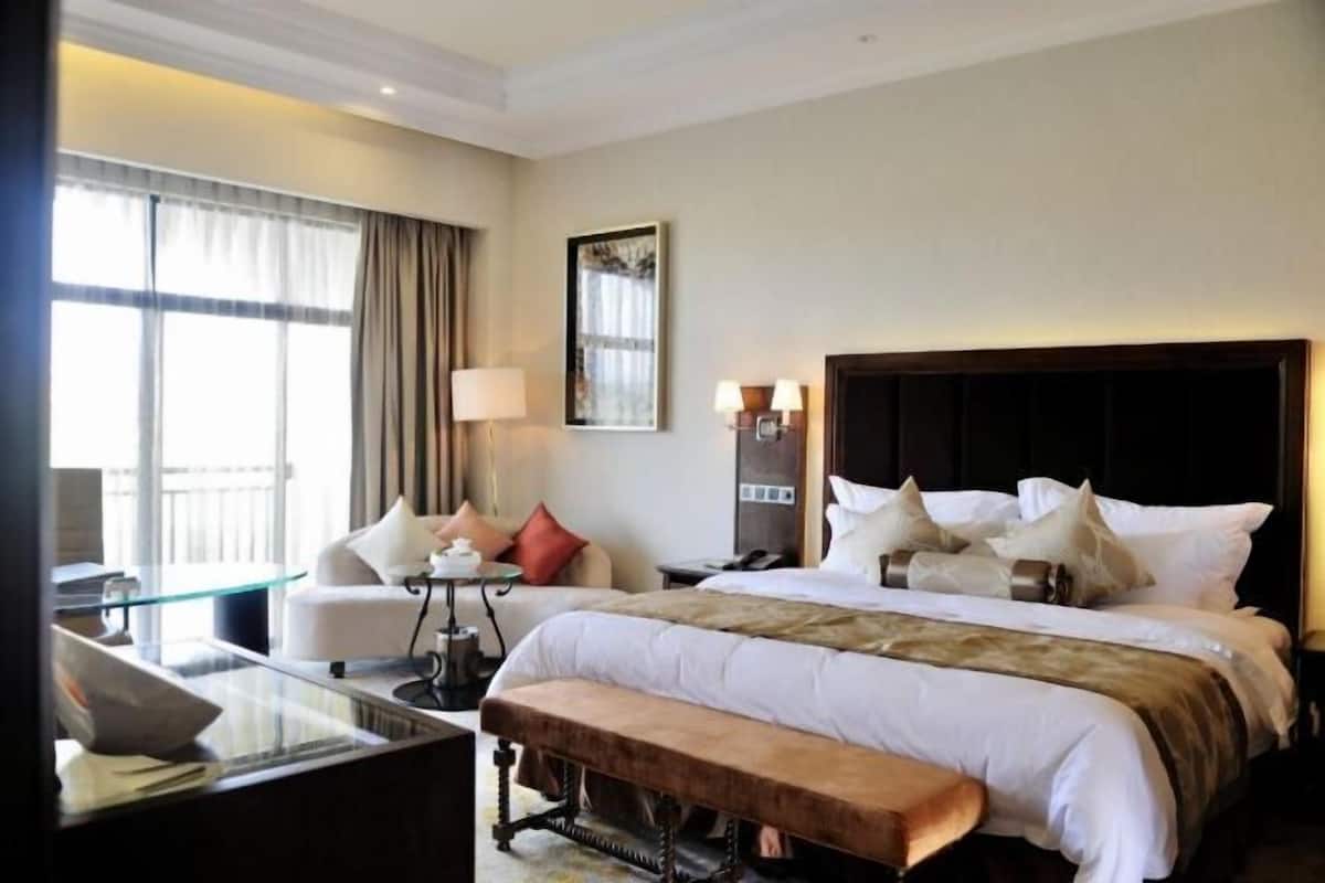A hotel room with a large bed, a desk with a chair, a bench, a window with curtains, and a wall-mounted lamp.