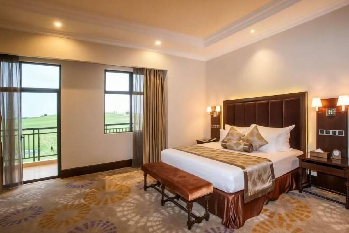 A hotel room with a large bed, a bench, a nightstand, and a view of a green landscape through the window.