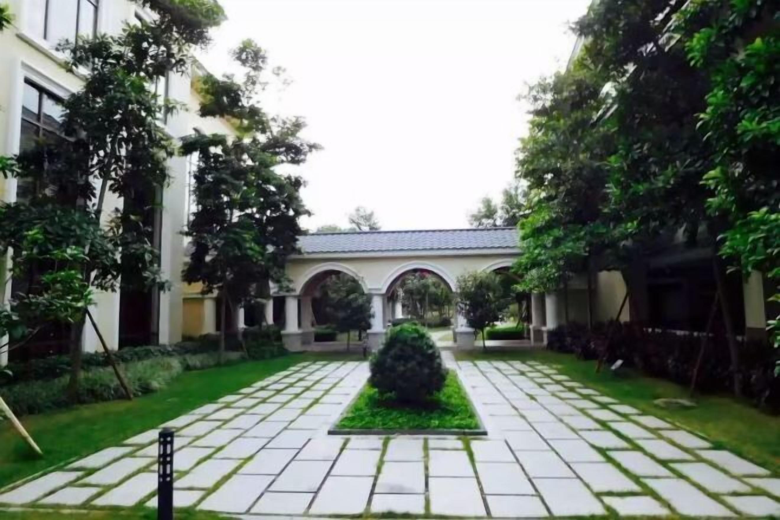 Une cour intérieure avec un chemin central, bordée de verdure et d’un bâtiment doté d’arcades.