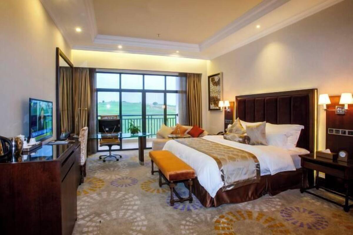 A hotel room with a large bed, a desk with a television, a balcony with a view, and a patterned carpet.