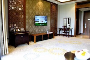 Un vestíbulo de hotel con un televisor montado en la pared, un banco de madera, un área de comedor con sillas y una alfombra estampada.
