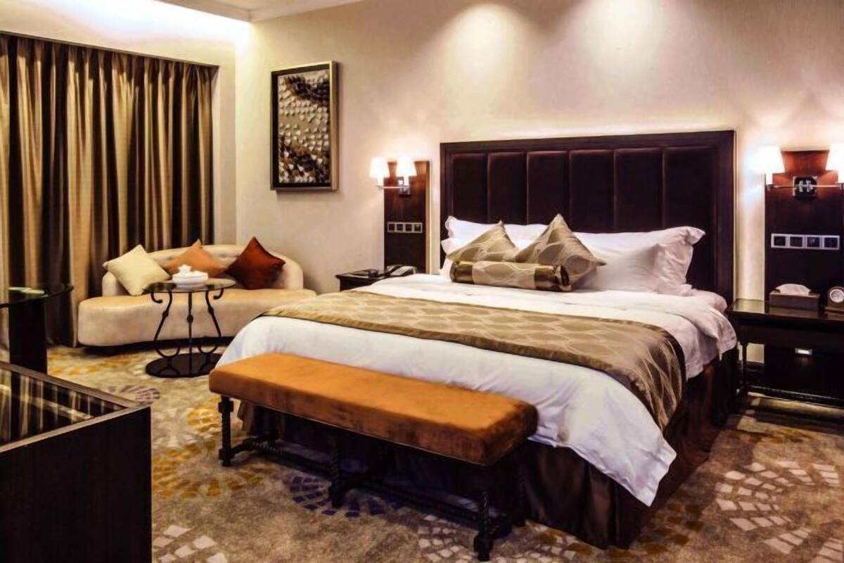 A hotel room with a large bed, a sofa, a bench, a small table, and a decorative wall piece.