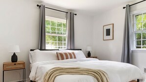 Een slaapkamer met een groot bed, twee nachtkastjes met lampen, een raam met gordijnen, en een lijst met een foto aan de muur.