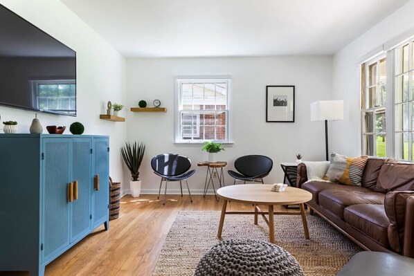 Una sala de estar moderna con un mueble azul, dos sillas negras, un sofá de cuero marrón, una mesa de centro redonda de madera y un ventanal con vistas a un edificio de ladrillo.