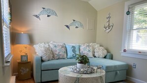 A cozy living room with a light blue sofa adorned with starfish pillows, a round coffee table with a leaf pattern, and a wall decorated with dolphin and anchor artwork. - Cozy Cape Cottage (Brewster)