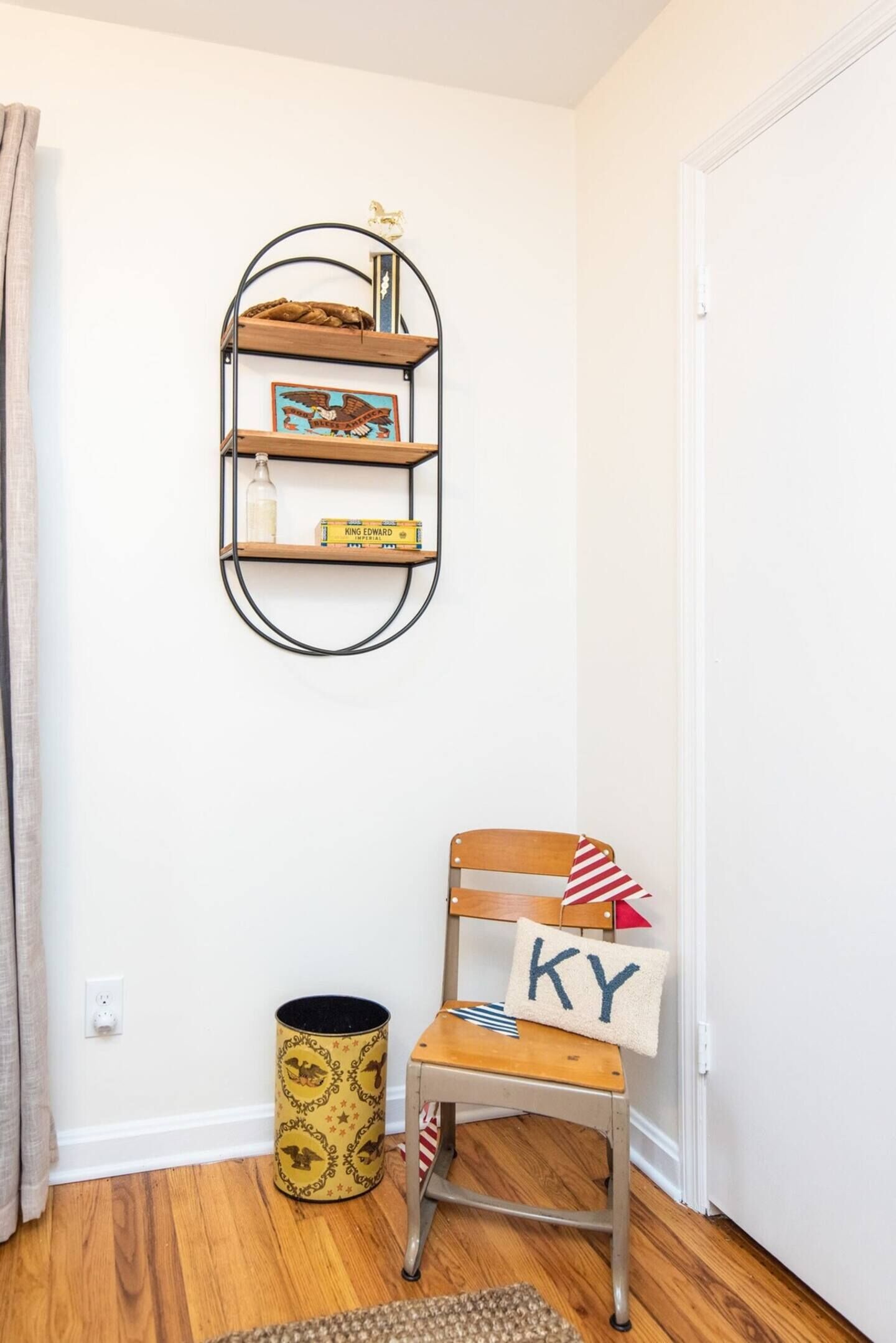Un coin avec une chaise en bois, une petite étagère avec des livres et une bouteille, et une poubelle.