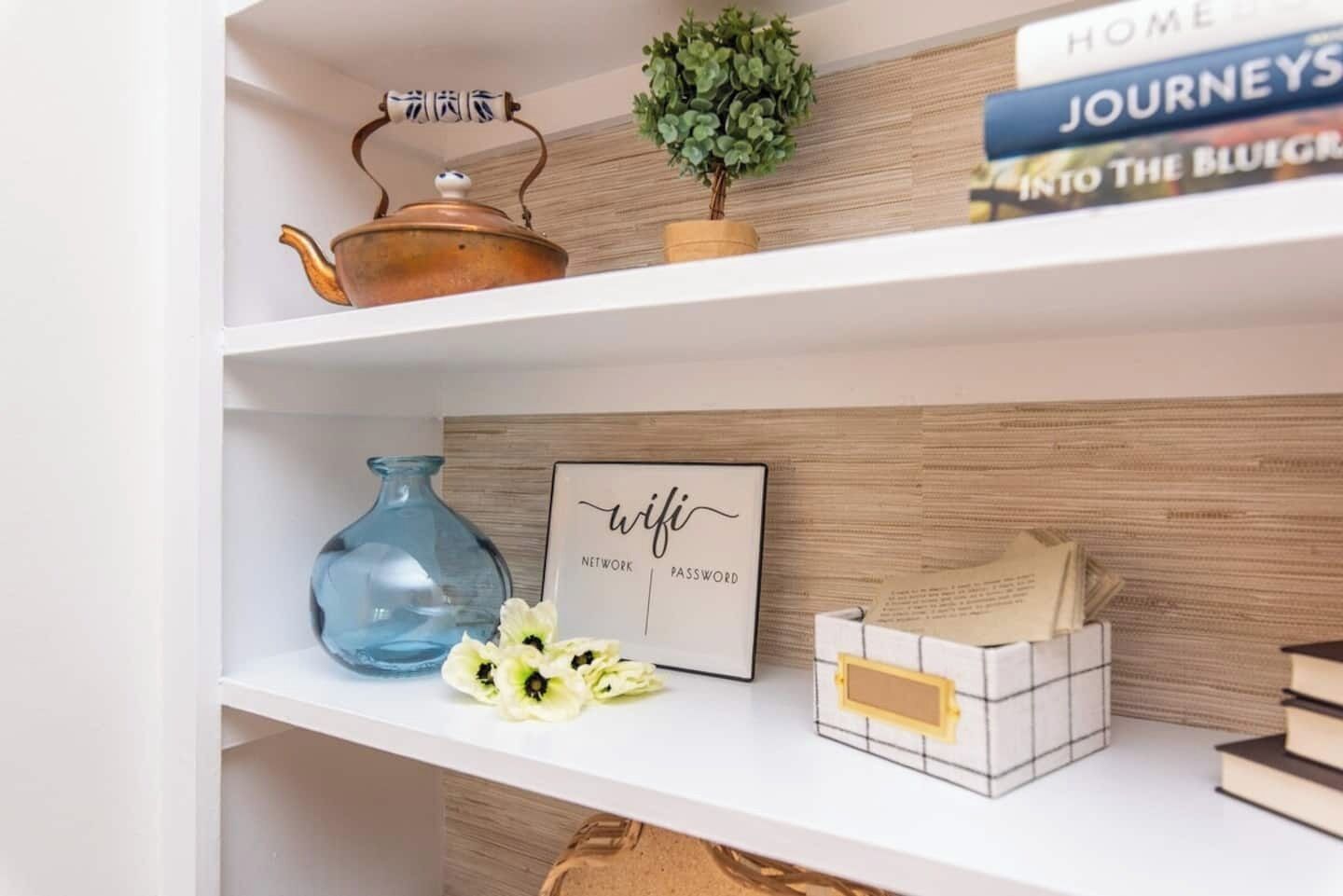A bookshelf with a teapot, a small plant, a framed sign, a vase with flowers, and a stack of books.