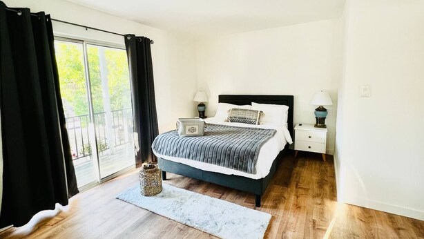 A bedroom with a bed, bedside tables, a window with curtains, a view of greenery outside, and a wooden floor.