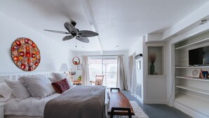 A bedroom with a bed, a bench, a ceiling fan, a large decorative wall piece, and a TV on a shelf.
