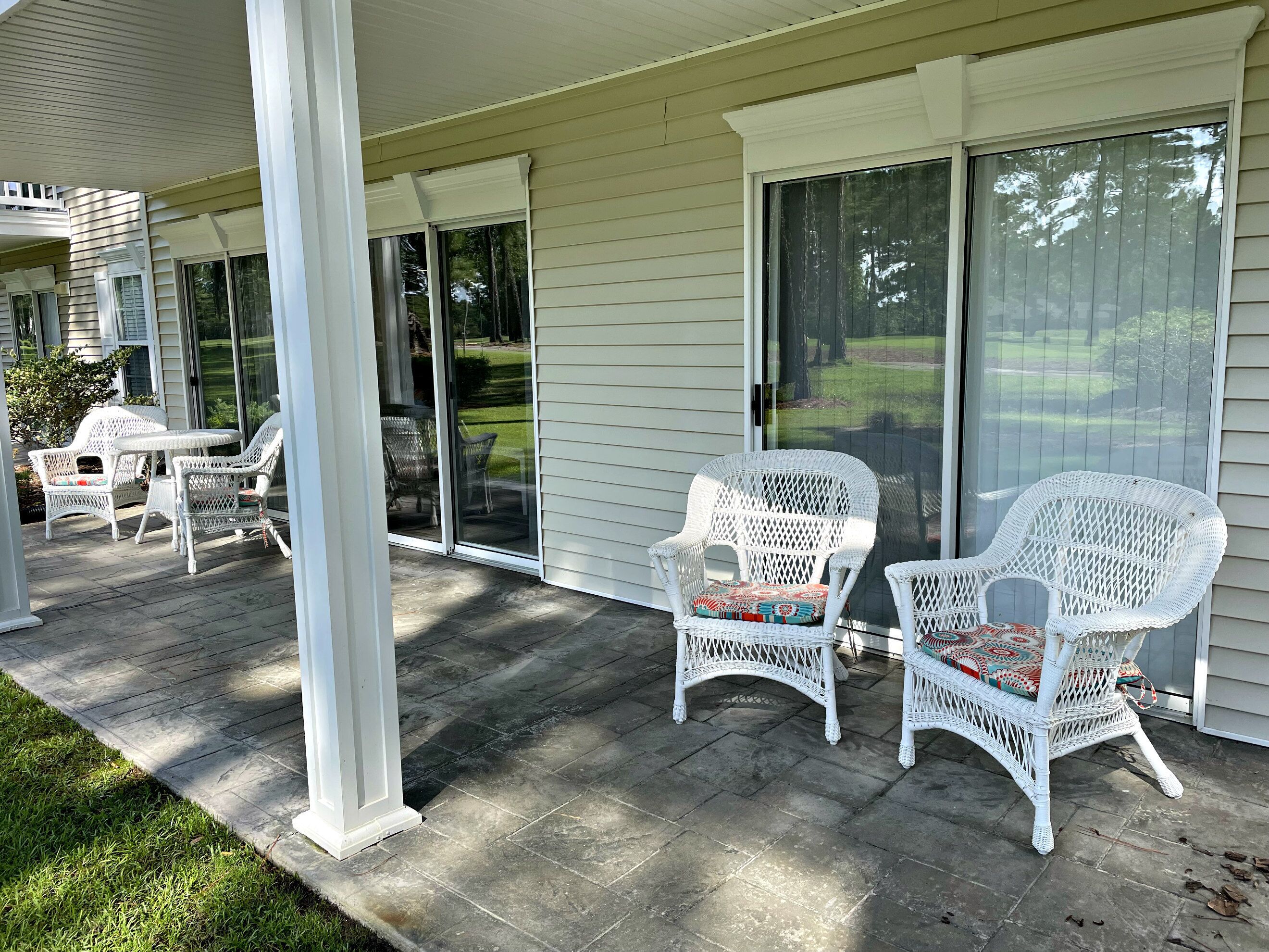 A covered patio with white wicker furniture, including chairs and a table, and a sliding glass door leading to a grassy yard.