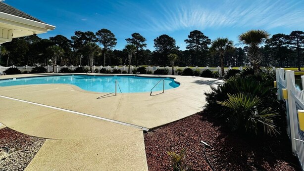 Une piscine avec une plage en béton, entourée d’une clôture blanche et d’arbres.