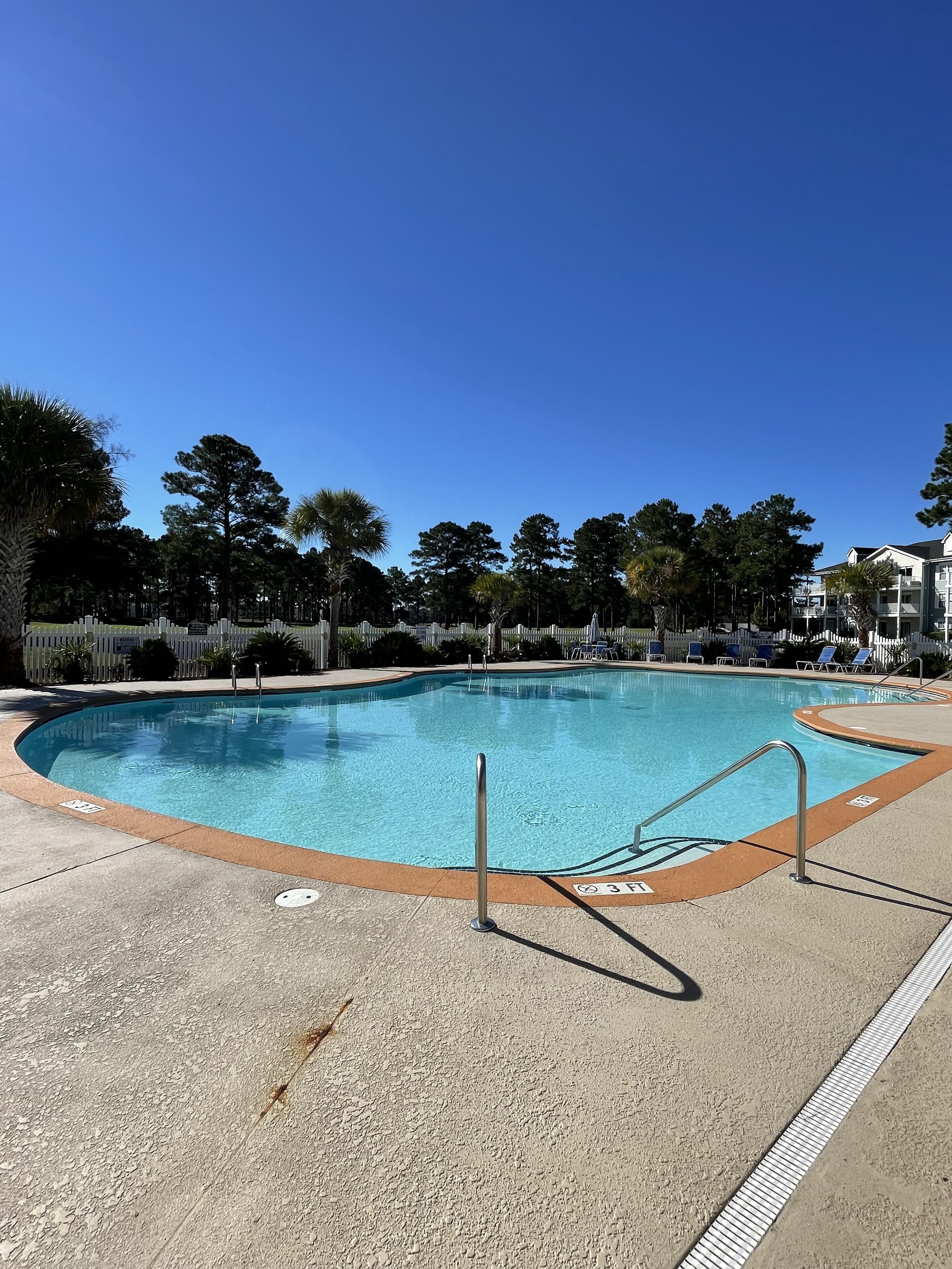 A clear blue pool with a surrounding deck, a fence, and trees in the background.