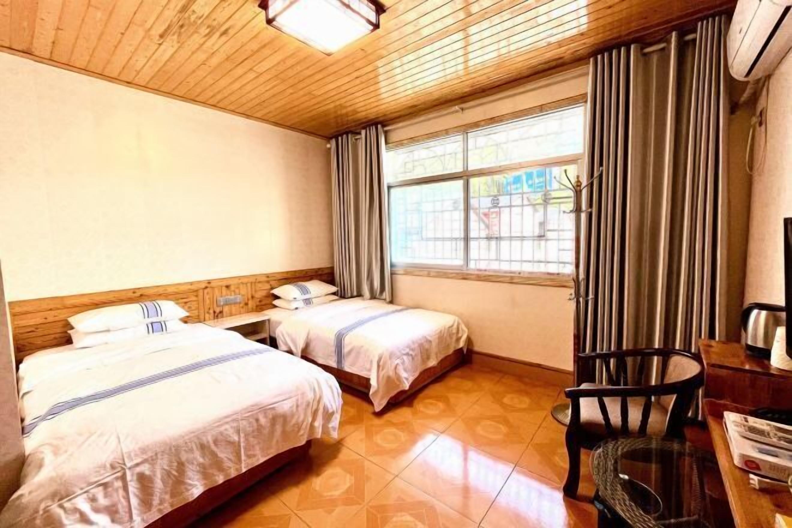 A hotel room with two beds, a wooden ceiling, a window with curtains, a chair, and a table with a kettle.