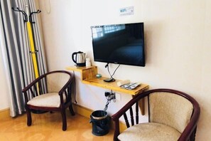 A room with a TV mounted on the wall, two wooden chairs, a small wooden table with a kettle, and a WiFi sign.