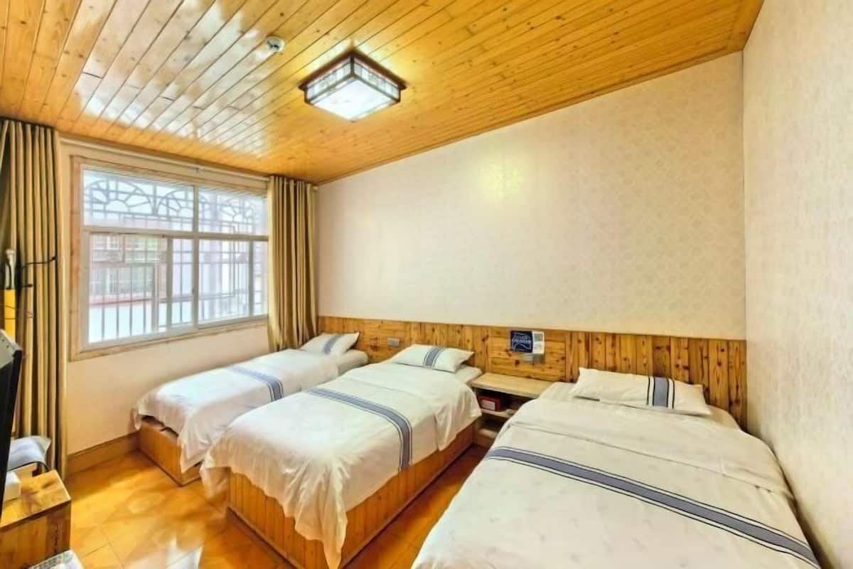 Ein Zimmer mit drei hölzernen Betten, einem fenster mit Vorhängen, einer Holzbalkendecke und einem kleinen Tisch.
