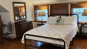 Une chambre avec un plancher en bois, un lit recouvert d’une couvre-lit à motifs floraux, une commode, une chaise, un bureau et une lampe.