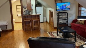 Un salon avec un canapé rouge, un fauteuil noir, un plancher en bois, un ventilateur de plafond et un téléviseur fixé au mur.