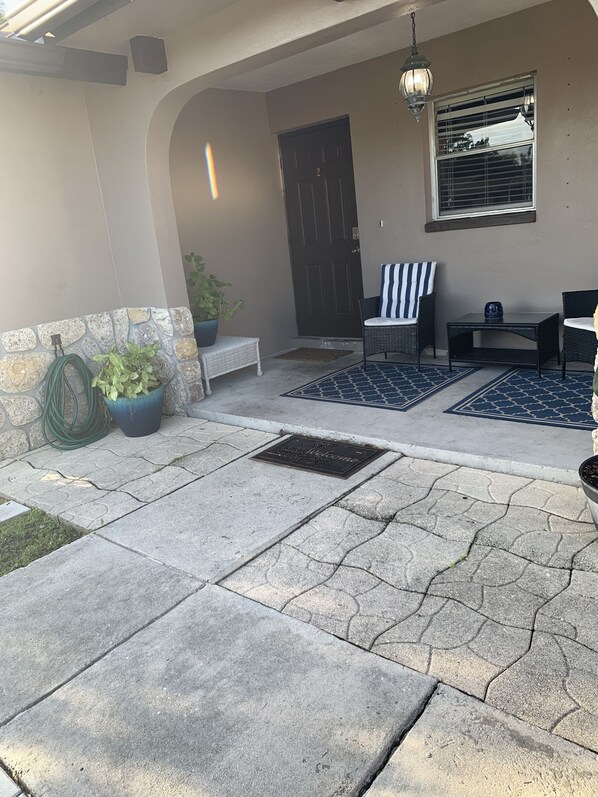 A covered patio area with stone flooring, a welcome mat, potted plants, outdoor furniture, and a door.