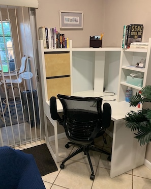 A corner office with a white desk, a black office chair, a bookshelf with various books, a framed picture on the wall, and a small plant on the desk.