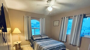 Ein Schlafzimmer mit einem Bett, einem Deckenventilator, einer Lampe, einem kleinen Tisch und einem Fenster mit Vorhängen.